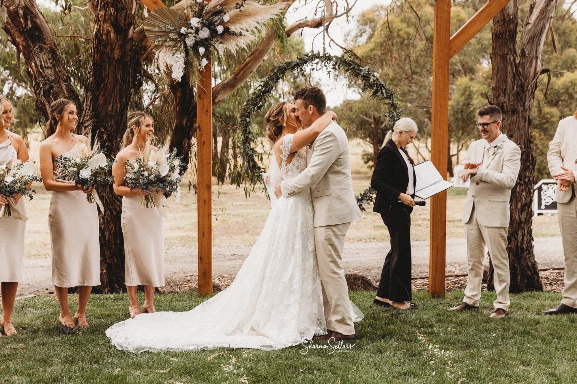 Ballarat wedding ceremony with floral backdrop hired from Bloomy Days. Bride and groom are kissing with wedding party watching.