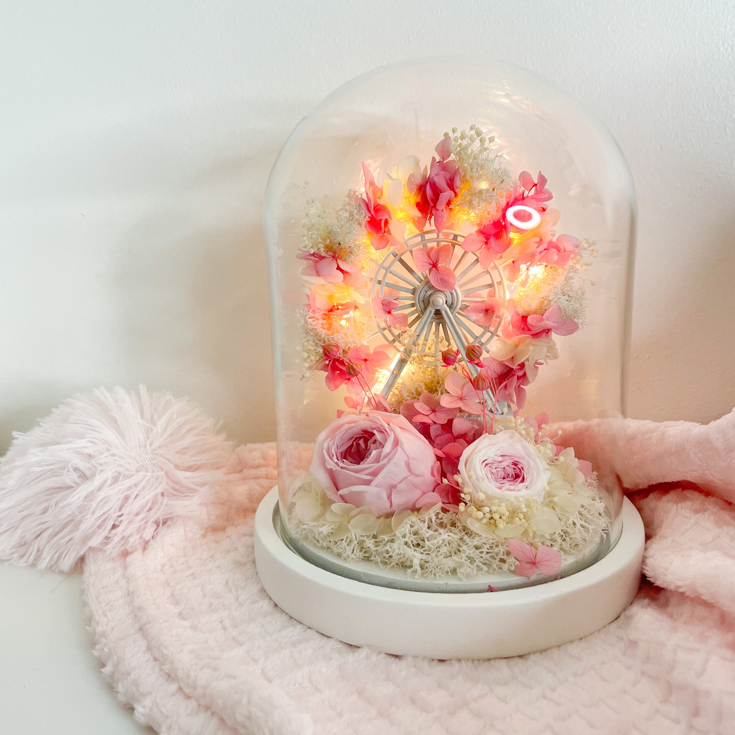 A glass dome featuring a pink floral arrangement with a ferris wheel and battery-powered lights inside, placed on a pink surface.